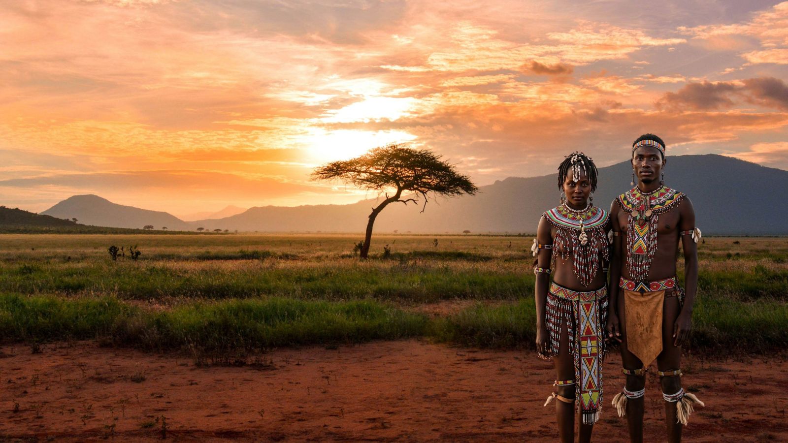 A Khoisan couple stands in an open field during an African sunset wife practices labia elongation, wearing traditional attire, with a tree silhouetted in the background.